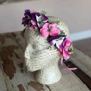 May include: A purple and pink flower crown with green leaves. The crown is made of artificial flowers and is sitting on a white mannequin head.