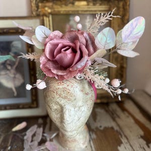 May include: A decorative headpiece featuring a large, dusty rose-colored rose and iridescent leaves. The headpiece is adorned with small, white berries and sits atop a head-shaped form covered in sheet music. A pink ribbon secures the piece.