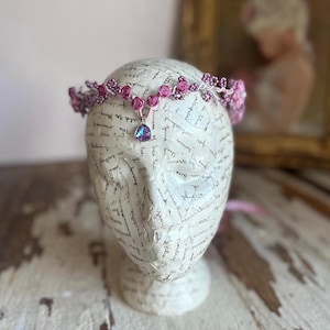 May include: A delicate pink and purple floral crown with a small iridescent gemstone pendant. The crown is made of small fabric roses and beads, and is displayed on a white mannequin head.