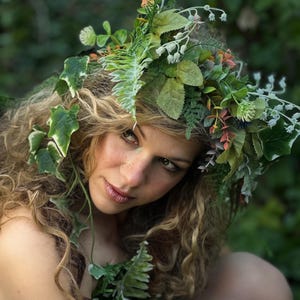 May include: A woman with curly blonde hair adorned with a vibrant floral crown of green leaves, white flowers, and orange accents. The woman is surrounded by lush green foliage, creating a natural, earthy aesthetic.