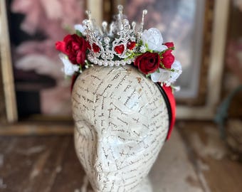 May include: A headband featuring a silver-toned metal crown adorned with small red heart-shaped gems and white pearls. The crown is flanked by artificial red and white roses and small green leaves, all mounted on a red band worn on a mannequin head.