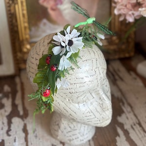 May include: A floral headpiece featuring white daisy-like flowers, green leaves, and red ladybugs. A green dragonfly is perched on top. The headpiece is displayed on a mannequin head with script-like text.