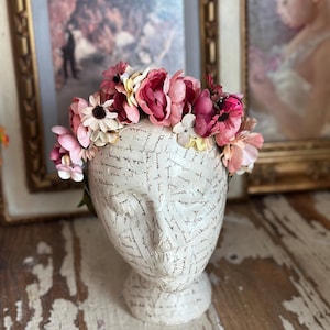 May include: A floral crown of pink and cream-colored flowers adorns a white papier-mâché head. The head is covered in script and sits on a pedestal. The background includes framed artwork and a distressed wooden surface.