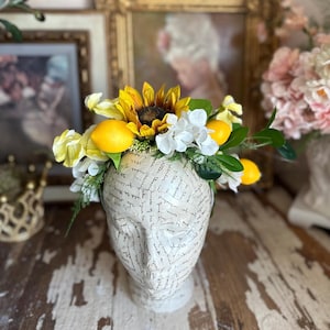 May include: A decorative head sculpture adorned with a vibrant floral arrangement. The arrangement features a large sunflower, yellow lemons, white flowers, and green foliage. The head is cream-colored with script writing.