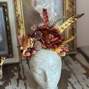 May include: A decorative headpiece featuring a mix of faux flowers, feathers, and foliage in shades of brown, orange, and gold. The headpiece includes a pinecone and faux berries. The headpiece is displayed on a white mannequin head with script writing.