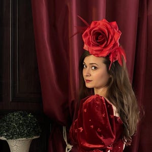 May include: A woman wearing a large red rose headpiece with red feathers. She is wearing a red velvet dress with butterfly accents. The background includes a dark wood door and a burgundy curtain.