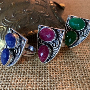 May include: Three silver rings with different colored gemstones. The rings are in the shape of a shield with ornate designs. The gemstones are blue, red, and green.