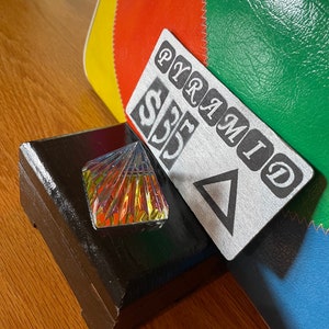 May include: A clear glass pyramid-shaped bottle stopper with a gold label on a dark brown wooden box. The box is sitting on a wooden surface. A white tag with the text "PYRAMID" and "$35" is leaning against the box.