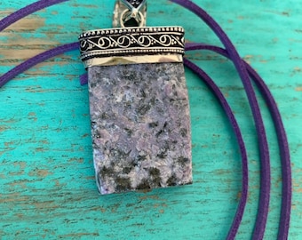 May include: A purple and grey stone pendant with a silver ornate frame and a purple suede cord.