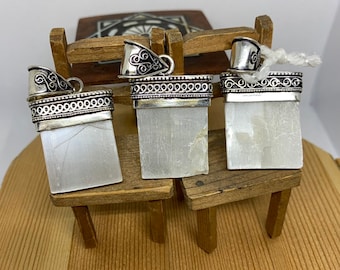 May include: Three silver pendants with white selenite stones. The pendants have intricate silver designs and are displayed on a wooden surface.