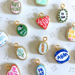 May include: A collection of small, hand-painted charms with gold-colored edges and loops. Each charm features a different design, including text like "Run into Jesus," "Amen," and "Seek Jesus," along with other religious symbols and phrases.