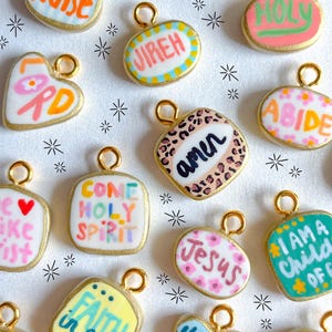 May include: A collection of colorful, hand-painted pendants with gold-colored edges and loops. Each pendant features a different word or phrase, such as "Praise," "Jireh," and "Jesus," in various fonts and colors. The pendants are set against a white background.