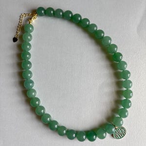 May include: A beaded necklace featuring smooth, round, light green beads strung together. The necklace includes a gold-toned metal extender chain with a small heart-shaped charm at the end and a round gold-toned pendant with the text WWJD in green.