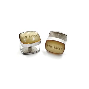 Può includere: Un paio di gemelli d'argento con una faccia rettangolare di colore giallo traslucido. La faccia di ogni gemello ha il testo "TED BAKER" inciso.