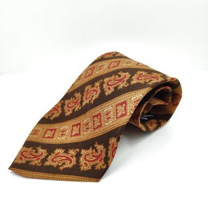 Può includere: Una cravatta marrone con un motivo paisley rosso e oro. La cravatta presenta strisce alternate di motivi paisley e geometrici. La cravatta è realizzata in tessuto liscio ed è adatta per occasioni formali.