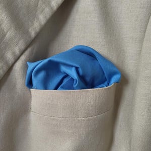 Peut inclure: Gros plan sur une veste en lin beige avec une pochette de costume bleu clair. La pochette est pliée et insérée dans la poche poitrine de la veste, créant un contraste de couleur avec le tissu neutre.