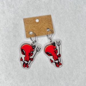 May include: A pair of devil-themed earrings. Each earring features a red devil cartoon character holding a pitchfork, with a white diaper and black horns. The earrings are attached to silver-colored hooks and a brown cardboard backing.
