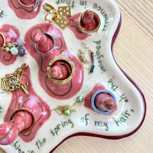 May include: A white ceramic ring holder with a wavy shape, featuring pink and green painted designs. The holder has several ring posts and holds various rings and jewelry. Text on the holder reads, "...in the spring of my hair, imagined romance..."