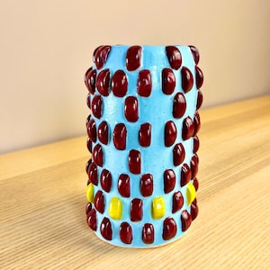 Puede incluir: Un jarrón cilíndrico con un fondo azul claro y un patrón de rectángulos elevados de color rojo oscuro y amarillo. El jarrón está sobre una superficie de madera.