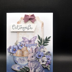 handgemachte Osterkarte mit Hase, Kaninchen, in Porzellantasse edle Eier, blau weiße Stiefmütterchen im Hintergrund, Vintagestil, Schleife