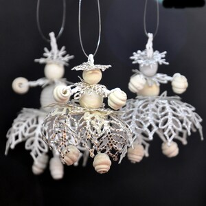 Könnte beinhalten: Drei handgefertigte weiße Muscheldoll-Ornamente hängen vor einem schwarzen Hintergrund. Jede Puppe hat einen Holzkopf, Muschelarme und einen dekorativen Rock. Die Puppen sind an dünnen, transparenten Schnüren aufgereiht.