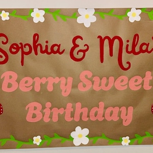 Puede incluir: Una pancarta de papel marr&oacute;n con el texto "Sophia & Mila's Berry Sweet Birthday" en rojo y rosa. La pancarta est&aacute; decorada con vides verdes, flores blancas y fresas rojas.