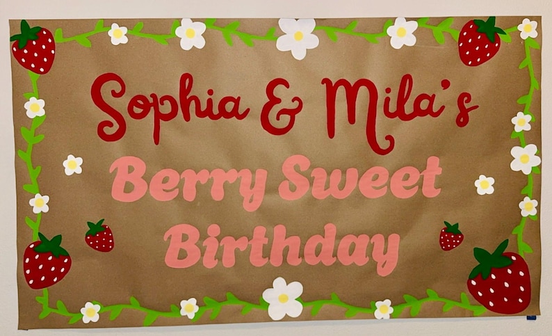 Puede incluir: Una pancarta de papel marr&oacute;n con el texto "Sophia & Mila's Berry Sweet Birthday" en rojo y rosa. La pancarta est&aacute; decorada con vides verdes, flores blancas y fresas rojas.