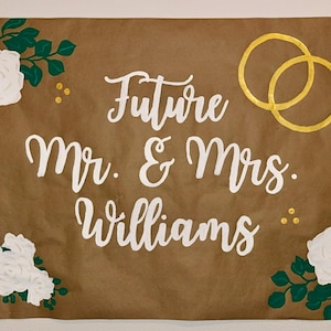 Puede incluir: Un letrero de papel marr&oacute;n con la inscripci&oacute;n blanca &laquo;Future Mr. & Mrs. Williams&raquo;. Anillos dorados y un anillo de diamantes en la parte superior. Rosas blancas y hojas verdes en las esquinas.