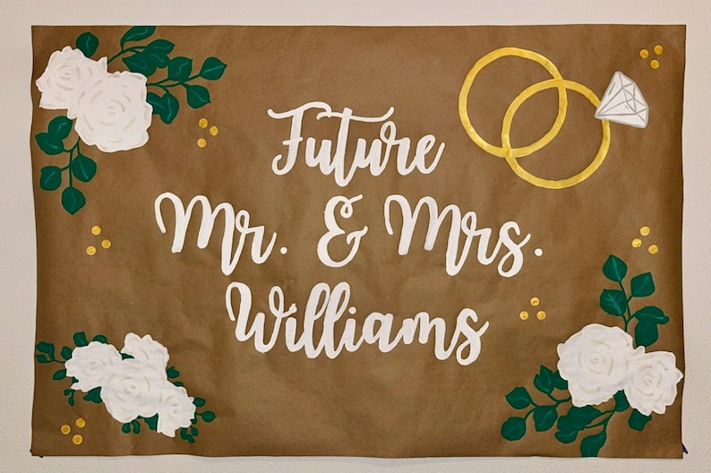 Puede incluir: Un letrero de papel marr&oacute;n con la inscripci&oacute;n blanca &laquo;Future Mr. & Mrs. Williams&raquo;. Anillos dorados y un anillo de diamantes en la parte superior. Rosas blancas y hojas verdes en las esquinas.