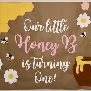 Puede incluir: Cartel de papel marr&oacute;n con el texto "Our little Honey B is turning One!" en escritura blanca y rosa. El cartel presenta panales de abeja amarillos, abejas, flores y un tarro de miel.