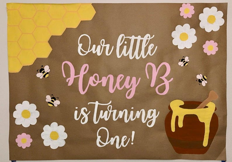 Puede incluir: Cartel de papel marr&oacute;n con el texto "Our little Honey B is turning One!" en escritura blanca y rosa. El cartel presenta panales de abeja amarillos, abejas, flores y un tarro de miel.