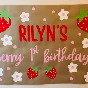 Puede incluir: Un letrero de papel marr&oacute;n con el texto "RILYN'S berry 1st birthday" en letras rojas y rosas. El letrero est&aacute; decorado con fresas rojas, flores blancas y lunares rosas. Una pancarta festiva de cumplea&ntilde;os.