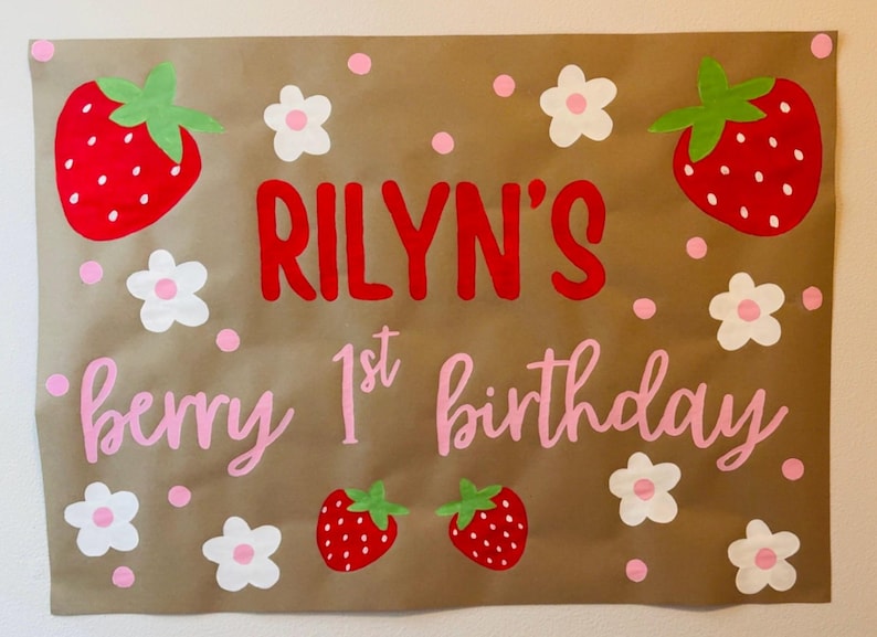 Puede incluir: Un letrero de papel marr&oacute;n con el texto "RILYN'S berry 1st birthday" en letras rojas y rosas. El letrero est&aacute; decorado con fresas rojas, flores blancas y lunares rosas. Una pancarta festiva de cumplea&ntilde;os.