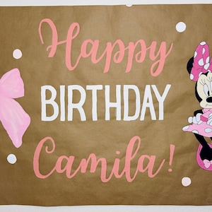 Puede incluir: Una pancarta de papel marr&oacute;n con las palabras "Happy BIRTHDAY Camila!" en rosa y blanco. Un lazo rosa y Minnie Mouse est&aacute;n en la pancarta. Lunares blancos est&aacute;n dispersos alrededor del texto y las im&aacute;genes.