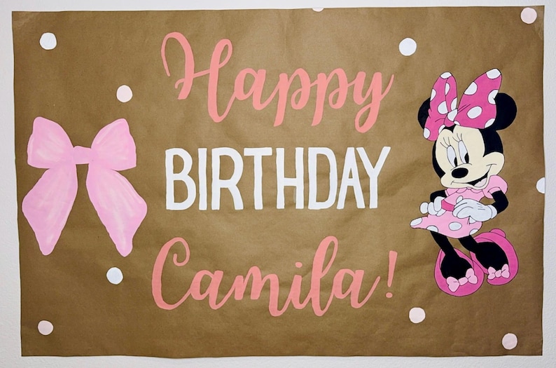 Puede incluir: Una pancarta de papel marr&oacute;n con las palabras "Happy BIRTHDAY Camila!" en rosa y blanco. Un lazo rosa y Minnie Mouse est&aacute;n en la pancarta. Lunares blancos est&aacute;n dispersos alrededor del texto y las im&aacute;genes.