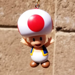 May include: A red and white mushroom character with a blue and yellow vest, a smiling face, and outstretched arms. The character is a popular video game character.