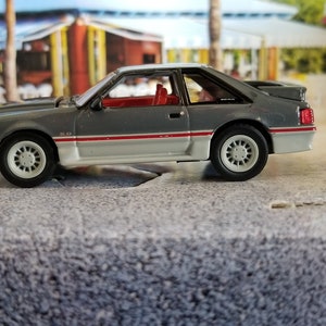 May include: A gray and white 1980s Ford Mustang model car with red accents. The car is a 1/18 scale model and is in excellent condition.