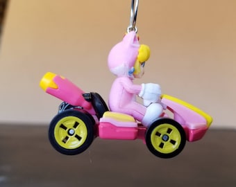 Könnte beinhalten: Ein rosa und gelbes Spielzeug-Rennwagen mit einer Figur eines rosa gekleideten Charakters am Steuer. Der Charakter hat blonde Haare und einen rosa Helm mit Katzenohren.