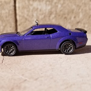 May include: A metallic purple toy car model of a Dodge Challenger coupe with a silver metal eyelet screw attached to the roof. The vehicle features dark gray multi-spoke wheels, a black rear spoiler, and clear windows showing a black interior cabin.