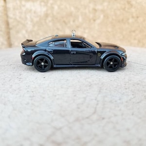 May include: Black die-cast toy car model of a Dodge Charger with black wheels. The car has a small silver ornament on the roof.