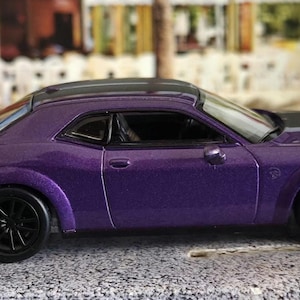 May include: A purple die-cast model car with black wheels and a black spoiler. The car has a black hood and roof. The vehicle is a coupe with a sleek, sporty design, and is set against a blurred background.