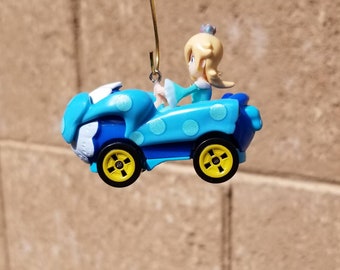 May include: A blue and white toy car with a polka dot design. A blonde haired figure with a crown sits in the driver's seat.