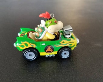 May include: A green toy car with yellow flames and a Bowser figure behind the wheel. The car has black wheels and a silver exhaust pipe.