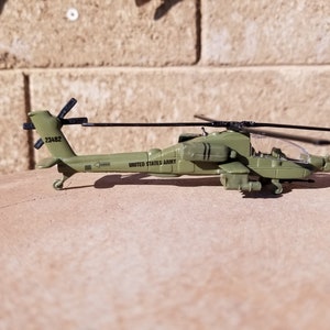 May include: A green toy helicopter with black rotors and the words "UNITED STATES ARMY" printed on the side. The model has the number "23482" on the tail and is set against a textured background.