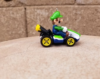 May include: A green and white toy race car with a green and black figure of Luigi driving. The car has a black exhaust pipe and yellow wheels.