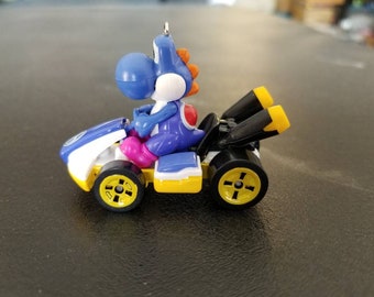 May include: A blue and yellow toy race car with a blue Yoshi character behind the wheel. The car has black exhaust pipes and yellow wheels.