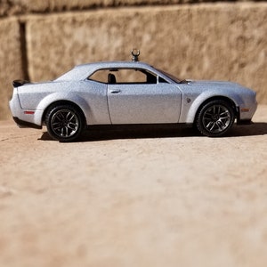 May include: A silver toy car with black wheels and a black spoiler. The car is a coupe with a detailed interior and is sitting on a light brown surface. The car has a small antenna on the roof.