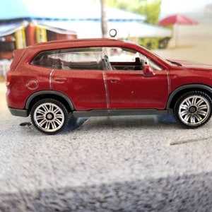 May include: A miniature red SUV toy with black tires and silver rims. The toy car has a detailed design, including windows and a roof rack. The background shows a beach scene with palm trees and a red umbrella.