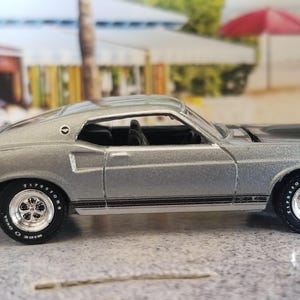 May include: Silver die-cast model of a classic Ford Mustang Mach 1. The car features black racing stripes, a rear spoiler, and Firestone tires. The model is set against a blurred background of a beach scene.