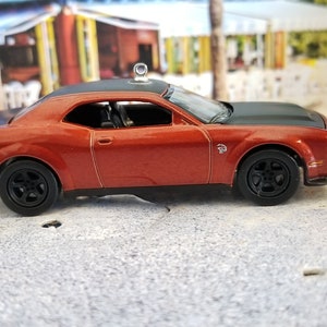 May include: A diecast model of a red and black muscle car with black wheels and a rear spoiler. The car has a black hood and roof, and a silver ornament on the roof. The car is on a gray surface.